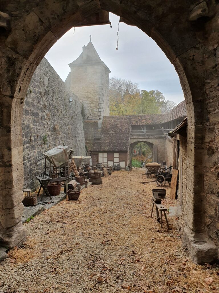 Mittelalterliche Szene. Blick durch ein Tor auf ein paar Stände links und Holzwägen rechts. Auf der Straße liegt Stroh und Laub.