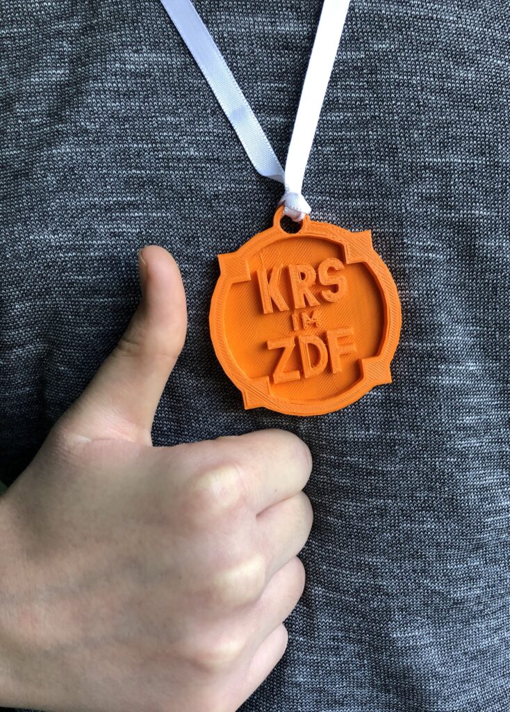 Eine mit einem 3D-Drucker gedrucktes Amulett auf dem "KRS im ZDF" steht. Daneben eine Hand mit Daumen nach oben.