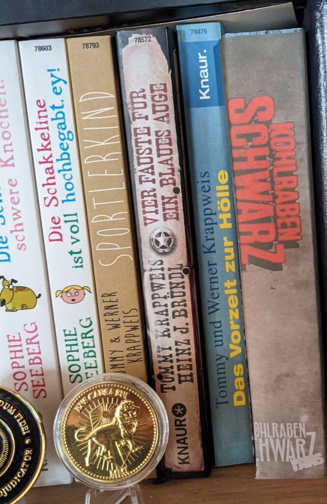 Verschiedene Buchrücken im Regal. Links sind zwei Bücher von Sophie Seeberg, dann Bücher von Tommy Sportlerkind, Vier Fäuste für ein blaues Auge, Das Vorzelt zur Hölle. Ganz rechts ist das Kohlrabenschwarz Buch mit einem gebasteltem Umschlag.