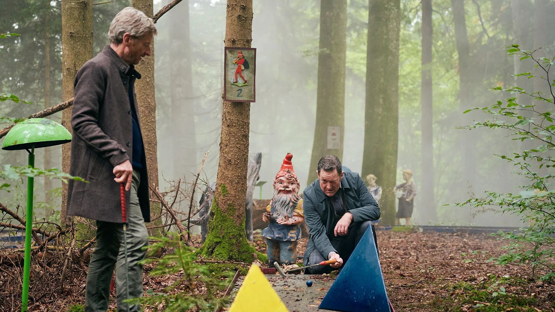 Stefan Schwab und Thomas Falbner spielen Minigolf in einem Wald. Thomas kniet neben einem Gartenzwerg und versucht die beste Bahn für seine Kugel zu finden.