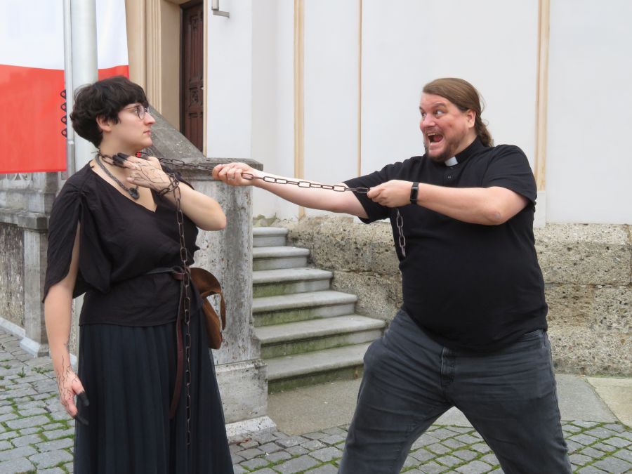 Linda als Perchta verkleidet hat einen Kette um den Hals. Ihre Fingerspitzen sind schwarz angemalt und sie berührt damit ihren Hals. Florian steht ihr als Franz gegenüber und zieht an der Kette