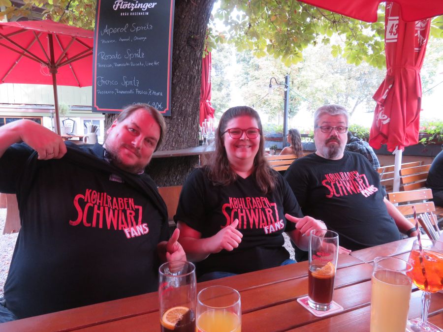 Im Biergarten sitzen Florian, Marina und Chris nebeneinander und haben ein schwarzes Shirt mit dem Logo der Kohlrabenschwarz Fans an.