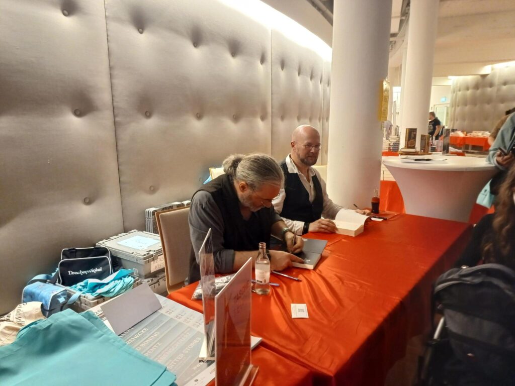 Tommy Krappweis und Christian von Aster sitzen am Tisch und geben Autogramme.